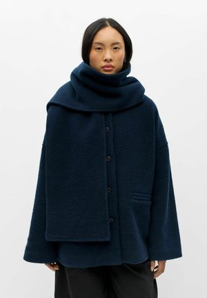 Femme portant un manteau oversize bleu foncé boutonné avec une grande écharpe assortie enroulée autour du cou, sur un fond uni.