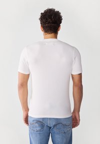 Hvit tettsittende t-skjorte med korte ermer, teksturert stoff, ribbestrikket halslinning og en jevn overflate. Bæres med lyseblå jeans med dekorativ brodering.