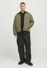 Olive green padded jacket with a zip front, worn over a black checkered shirt, paired with loose black cargo pants and black shoes.