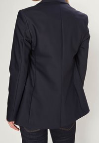 Tummansininen bleiseri, jossa on muotoiltu leikkaus, sileä kangas, rakenteelliset hartiat ja yksi takasauma. Yhdistetty tummansinisille farkuille.