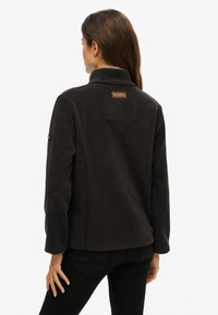 Veste en polaire noire avec un col haut et une plaque en cuir au dos. Présente une texture douce et des lignes simples et épurées.