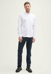 Camicia bianca a maniche lunghe con colletto alla coreana, abbinata a jeans blu scuro e scarpe nere. La camicia presenta una singola tasca sul petto.
