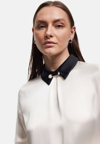 Blusa crema con cola de satén negro, que presenta un cierre decorativo con acentos de perlas; textura suave y detalle de frente plisado.