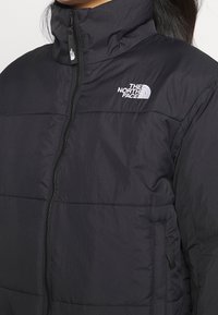 Black puffer jacket featuring a high collar, zip closure, and horizontal stitching. The North Face logo is visible on the left chest.
