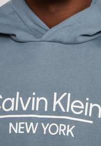 Felpa con cappuccio di colore azzurro chiaro realizzata in tessuto morbido, con testo bianco che recita "Calvin Klein NEW YORK" in un design curvo.