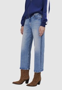 Jeans en denim bleu clair à jambes larges avec un effet délavé, assortis à des bottines en daim marron avec broderies florales et un talon bloc haut.