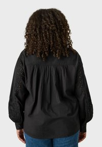Blusa nera con dettagli in pizzo intricati sulle maniche, schiena arricciata e polsini con bottoni, realizzata in un tessuto leggero.
