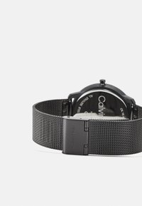 Svart mesh-rem klokke med rund klokkehus. Teksturert mesh-rem har et sikkert lås. Klar bakside med merkevarenavn synlig inni.