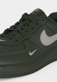 Σκούρο πράσινο αθλητικό παπούτσι Nike Air Force 1 με μαύρα κορδόνια και λευκά λογότυπα Nike swoosh στο πλάι και κοντά στην περιοχή των δαχτύλων.