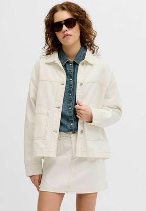 Kobieta z kręconymi włosami, w czarnych okularach przeciwsłonecznych, kremowej kurtce i spódnicy oraz niebieskiej dżinsowej koszuli zapinanej na guziki na jednolitym tle.