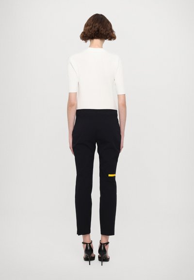 Pantaloni negri con croială, cu un buzunar de accent galben, asortați cu un top alb ajustat și pantofi cu toc înalt negri, văzuți din spate.