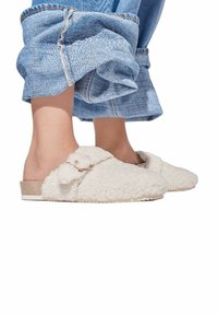 White furry slip-on slippers with a textured finish and a bow detail. Paired with loose denim pants showcasing rolled cuffs.