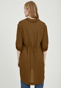 Robe chemise en tissu léger marron avec col, poignets à boutons, taille élastique et un ourlet arrière légèrement plus long. Finition texturée.