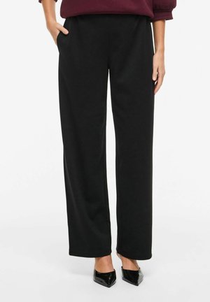 Osoba v voľných čiernych nohaviciach s rukou v vrecku, obutá do čiernych lodičiek so špicatou špičkou, stojaca na bielom pozadí.