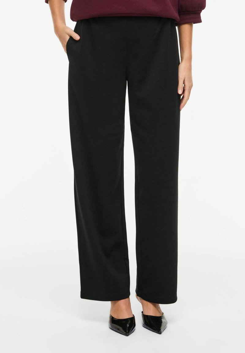 Henkilö, joka pukeutuu löysiin mustiin housuihin, käsi taskussa, yhdistettynä mustiin, teräväkärkisiin korkokenkiin, seisoo valkoisella taustalla.