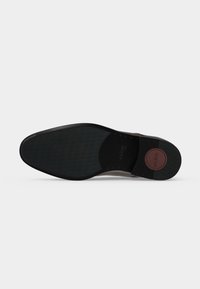 Semelle noire d’une chaussure habillée avec une prise texturée, une tache ronde marron près du talon estampillée "ALDO," et de petits clous le long de la zone de la voûte plantaire.