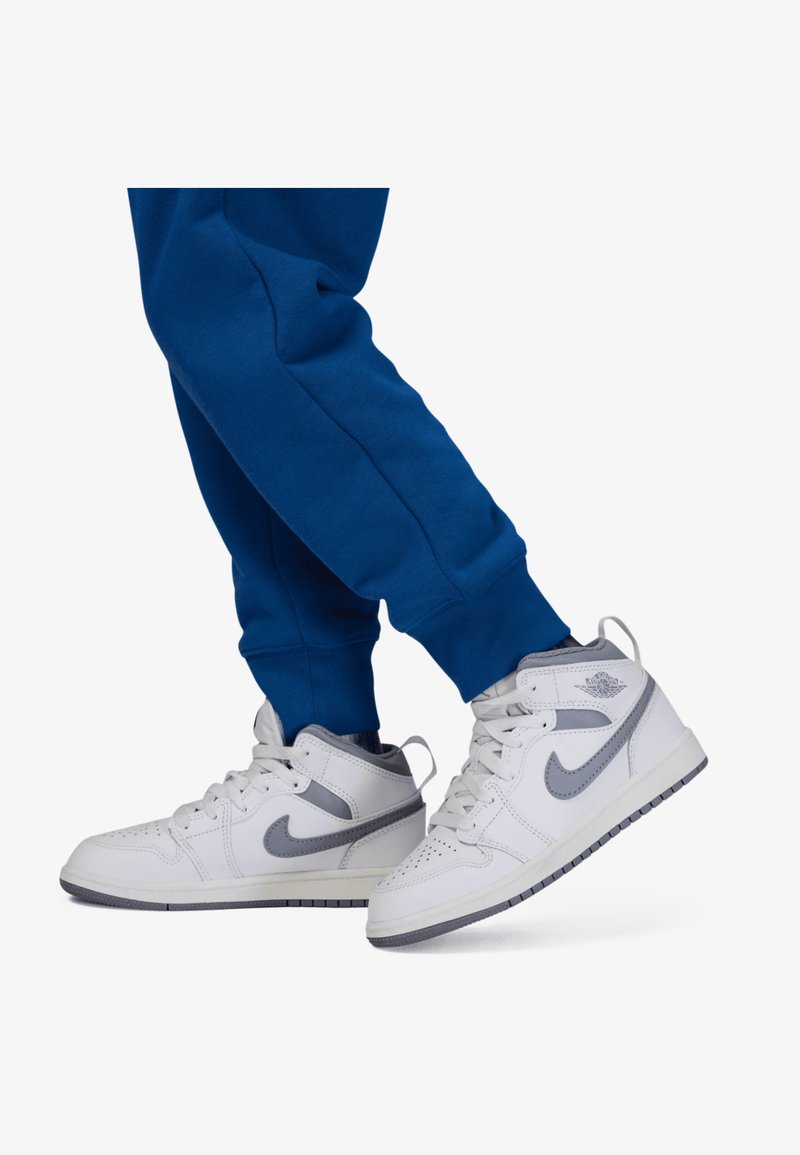 Zapatillas altas blancas con acentos grises y caja de puntera perforada, combinadas con pantalones de chándal azules que presentan puños elásticos y un diseño ajustado.