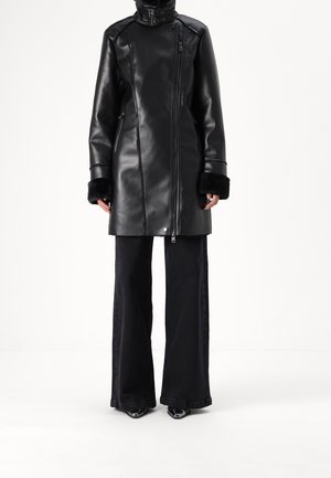 Persona con un abrigo de cuero negro con puños de piel, combinado con pantalones negros de pierna ancha y zapatos puntiagudos, de pie frente a un fondo blanco.