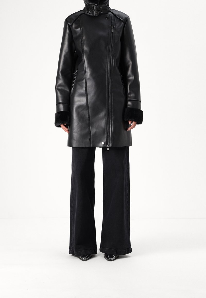 Personne portant un manteau en cuir noir avec des poignets en fourrure, associé à un pantalon noir à jambes larges et des chaussures pointues, debout devant un fond blanc.