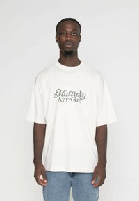 Bílá bavlněná tričko s oversize střihem, na přední straně s zeleným kurzívním nápisem "Multiply Apparel". Nosené s modrými denimovými džíny.