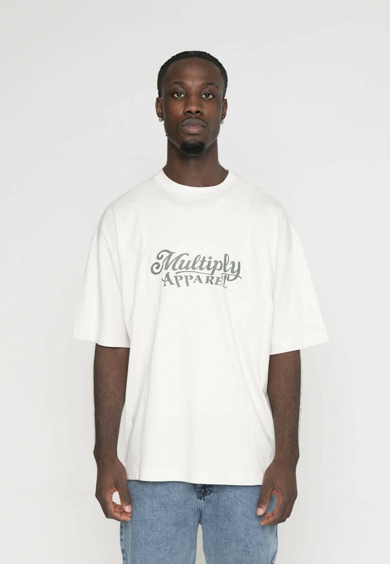 Bílá bavlněná tričko s oversize střihem, na přední straně s zeleným kurzívním nápisem "Multiply Apparel". Nosené s modrými denimovými džíny.