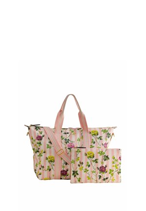 Kvinde iført en lyserød blomsterprintet langærmet skjorte og brede blomsterbukser, der holder en stor lyserød stribet blomstertragtaske, står op ad en blå væg.