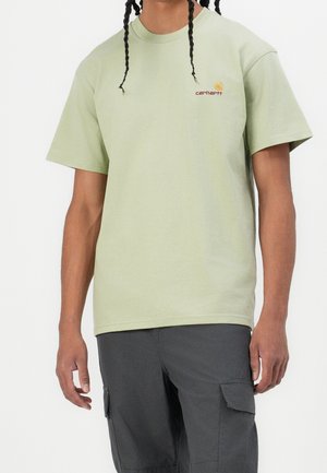 Person iført en lysegrøn Carhartt t-shirt og mørkegrå cargobukser, stående med armene afslappet ned langs siden.