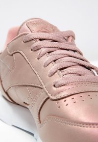 Gros plan sur une chaussure de sport en cuir rose clair avec des lacets assortis et des coutures détaillées sur un fond blanc.