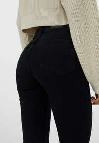 Jean noir taille haute avec une coupe ajustée, présentant un style classique à cinq poches, accompagné d'un pull en maille texturée de couleur beige clair porté au-dessus.