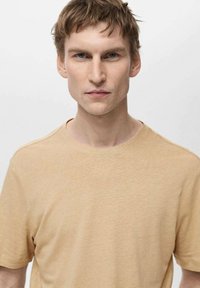 Tricou din bumbac bej, cu mânecă scurtă și guler rotund, cu un finisaj moale și texturat, expus pe un model pe un fundal neutru.