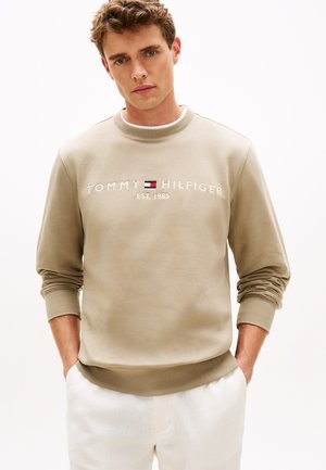 Noor mees, kellel on lühikesed lokkis juuksed, kannab beeži Tommy Hilfigeri logoga dressipluusi ja valgeid pükse, seisab käed taskutes.