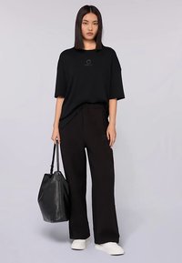 Svart oversize t-shirt med en liten logotyp, kombinerad med löst sittande svarta byxor. Modellen bär en svart läderhandväska och vita sneakers.