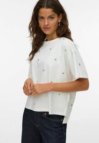 Hvid kortærmet top med små guldfarvede bjørnebroderier, en afslappet pasform og sideslidser. Parret med mørk denimjeans.