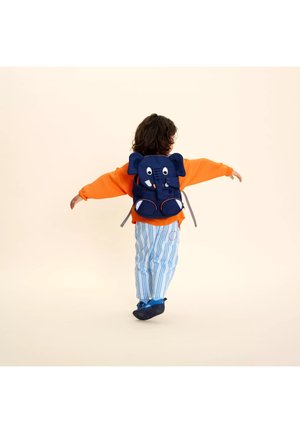 Child wearing orange sweater and blue striped pants walks with arms outstretched, carrying navy blue backpack shaped like an elephant.