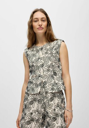 Femme portant un haut beige sans manches et un short assorti avec une broderie en spirale abstraite noire, debout devant un fond blanc uni.