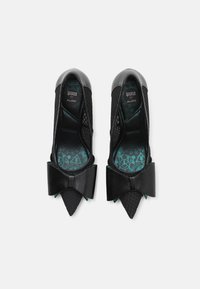 Ballerines synthétiques noires avec un bout pointu, des côtés en mesh et un détail de nœud proéminent. L'intérieur possède une semelle intérieure à motif turquoise.