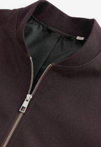 Veste marron foncé avec une fermeture éclair argentée à l'avant et un col côtelé, partiellement ouverte pour révéler une doublure intérieure noire texturée.
