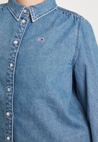 Camisa de mezclilla de botones con un pequeño logo rectangular en rojo, blanco y azul en el pecho, que presenta un cuello puntiagudo y una costura de hombro abullonada.