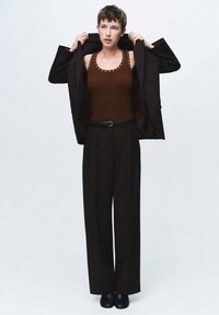 Top bej cu mânecă fără, accesorizat cu tinte, sacou negru, pantaloni largi și pantofi masivi negri, pe un fundal deschis.