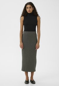 Žena s dugom kovrčavom kosom, obučena u crnu bezrukavnu turtleneck bluzu, sivu midi olovku suknju i crne baletanke, stoji naslonjena na bijelu pozadinu.