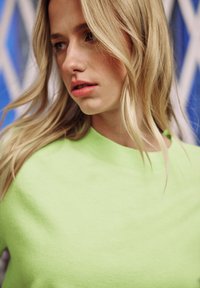 Maglione leggero verde neon con scollo tondo. Texture morbida e silhouette aderente, indossato con onde morbide e casual nei capelli.