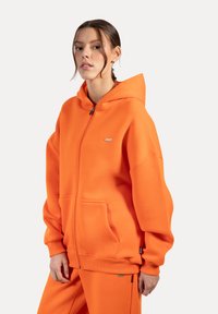Orange zip-up hoodie lavet af blødt materiale, med hætte, forlommer og ribbede manchetter. Lille logo detalje på brystet.