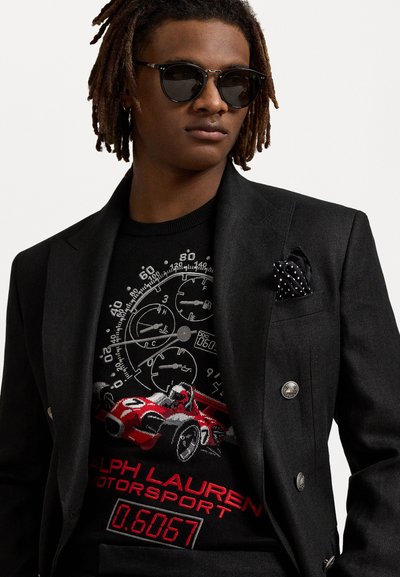 Blazer noir avec des boutons argentés sur un pull noir orné de graphiques de voitures de course rouges et de compteurs. Accentué par un mouchoir de poche noir et blanc à pois.