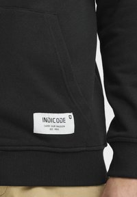 Haut à capuche noir avec une poche frontale, des poignets côtelés et un patch étiqueté blanc avec du texte. Tissu doux avec une texture lisse.