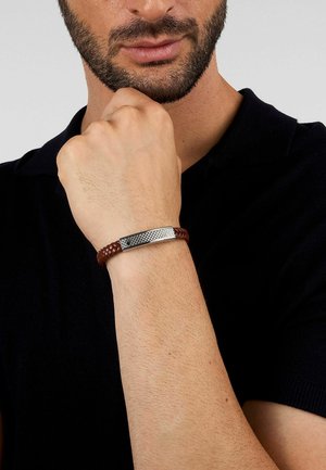 Mand med skæg iført en sort skjorte viser et brunt flettet læderarmbånd med tekstureret sølvspænde på hævet håndled nær ansigtet.