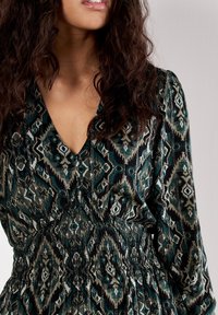 Long-sleeved dress with a deep V-neck, featuring a teal and black tribal pattern, gathered waist, and soft fabric texture.