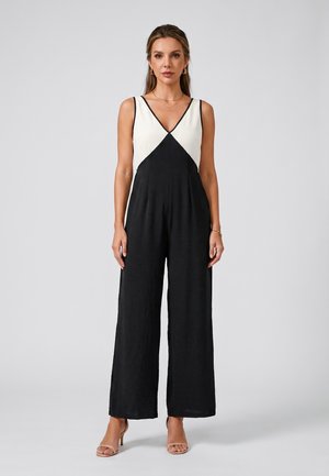Woman standing wearing sleeveless black wide-leg jumpsuit with white triangular top section and nude heels, neutral background.