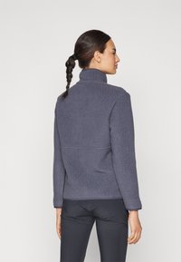 Giacca grigia in fleece con colletto alto e superficie testurizzata. Presenta maniche lunghe e un dettaglio sulla cucitura posteriore, indossata sopra pantaloni neri.