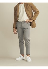 Mies, joka on pukeutunut vaaleanruskeaan nahkatakkiin, valkoiseen paitaan, harmaisiin housuihin ja valkoisiin lenkkareihin, seisoo yhdellä kädellä lantiolla vaalean beigeä taustaa vasten.