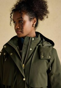 Oliwkowa wodoodporna kurtka z wysokim kołnierzem, złotymi detalami, zamek z przodu, podwójne kieszenie z klapami oraz logo na suwaku.